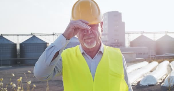 Retrato de ingeniero industrial sénior en gafas y chaleco salvavidas quitándose el casco de pie junto a una gran fábrica industrial y mirando a la cámara con luces solares. Concepto de estudio de industria e ingeniería . — Vídeo de stock