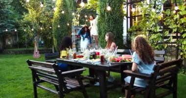 Afro-Amerikalı ve beyaz güzel kızlar bahçede yemek masasında oturuyorlar. Neşeli erkek ve kadınlar bahçeye giriyor ve arkadaşlarına sarılıyorlar. Asyalı kız ellerini sallıyor. Kutlama konsepti