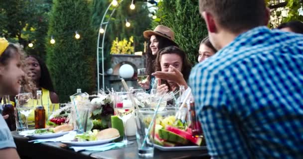 Multi-etnische jongeren zitten aan tafel en vermaken zich samen in de tuin. Afro-Amerikaanse en Kaukasische mooie meisjes glimlachen buiten. Vreugdevolle mannen glimlachend en pratend. Zomer concept — Stockvideo