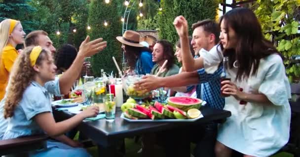 Joyeux jeunes gens multi-ethniques ensemble à la garden party. Les filles afro-américaines et caucasiennes rient. Des hommes et des femmes joyeux passant plat tout en bavardant. Jolie fille asiatique souriante. Concept d'été — Video