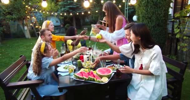 Jongeren eten en drinken aan tafel in de tuin. Afro-Amerikaans meisje dat lacht en drinkt. Blanke mannen die eten doorgeven. Aziatische vrouw lacht. Vrienden klinkende glazen. Partijconcept — Stockvideo