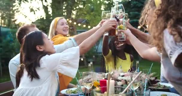 즐거운 혼혈의 젊은 친구들이 안경을 들고 윙크하는 모습. 코카서스 남자들 , 아시아 계 및 아프리카 계 미국인 소녀들이 정원에 있는 식탁에 함께 모여 이야기를 나누었다. 여름의 재미 컨셉 — 비디오