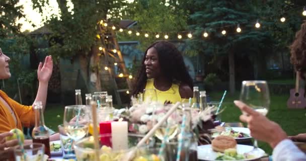 Veel multi-etnische jonge mannen en vrouwen zitten aan tafel in de tuin. Afro-Amerikaans meisje lacht. Blanke vrolijke jongens en meisjes knuffelen. Mensen verzamelen zich. Feestconcept — Stockvideo