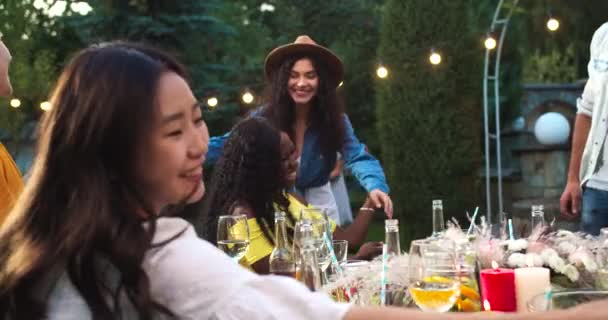许多混血的年轻快乐女人坐在花园的餐桌旁。白人女性拥抱非裔美国女孩。快乐的亚洲女孩在户外微笑。朋友们在一起聊天快乐的概念 — 图库视频影像