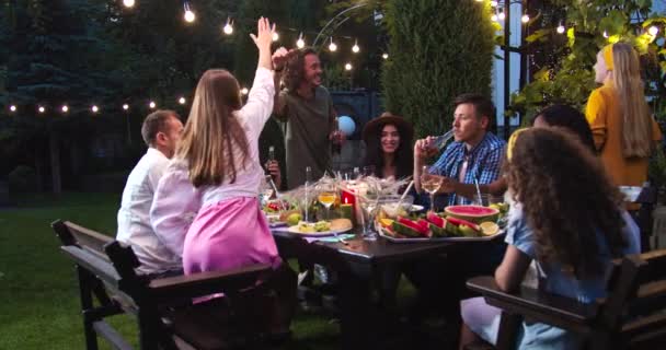 Gelukkige gemengde rassen jonge mensen spreken buiten op tuinfeest. Mooie blanke en Afro-Amerikaanse meisjes en mannetjes zitten aan tafel. Aziatische vrouw lacht. Mensen praten en heffen glazen en flessen — Stockvideo