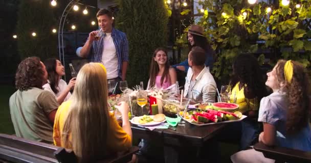 Vidám vegyes fajú barátok a kerti partin esténként. Kaukázusi fiatal férfiak és nők isznak és beszélgetnek a szabadban. Afrikai amerikai lány mosolyog. Ázsiai lány jó hangulatban. Pohárköszöntőt mondani — Stock videók
