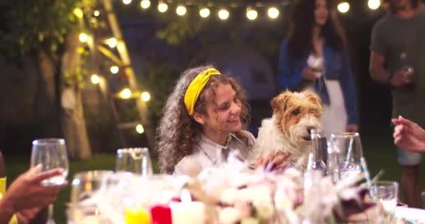 Молода кавказька красуня сидить за столом і тримає милого собаку. Багатоетнічні друзі веселяться на вечірці в саду вночі. Самці і самиці піднімають окуляри і кліпають. Дивна концепція — стокове відео
