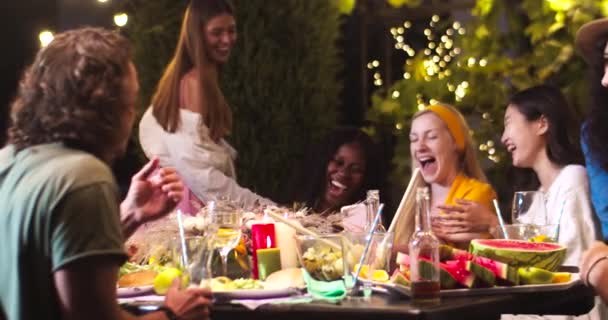 Joyeuses métissées jeunes femmes riant en s'asseyant à table à la fête en plein air et regardant smartphone. Les filles afro-américaines et asiatiques rient. Caucasiens beaux hommes parlant à la fête de jardin — Video