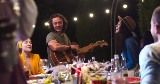 Radosna mieszana rasa młodych ludzi bawiących się wieczorem na imprezie w ogrodzie. Szczęśliwy biały mężczyzna gra na gitarze i śpiewa na świeżym powietrzu. Ładna Afroamerykanka z uśmiechem na twarzy. Koncepcja czasu wolnego — Wideo stockowe