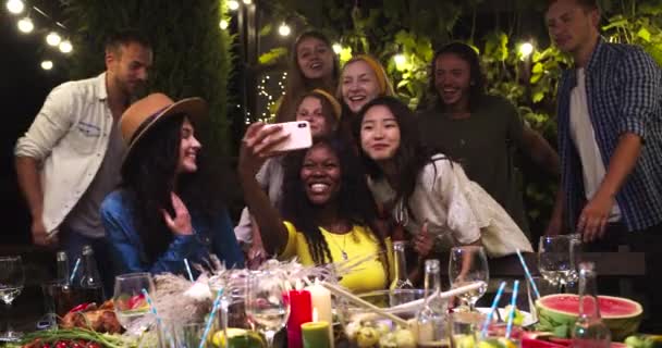 Різні багатонаціональні молоді люди зібралися разом на городній вечірці. Афроамериканська дівчина сидить і робить фотографії на мобільному телефоні. Азійська дівчина махає рукою. Привабливі кавказькі хлопці посміхаються — стокове відео