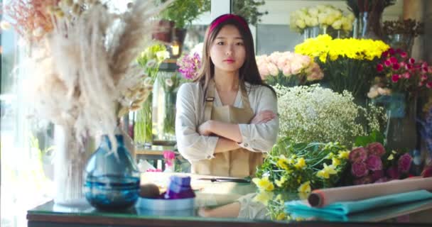 Een knappe jonge vrouw met een schort, werkend in een bloemenwinkel. Aantrekkelijke Aziatische vrouwelijke bloemist kruisen armen en kijken naar de camera. Bloemenwinkel, bloesems, kamerplanten, beroep, bedrijf. — Stockvideo