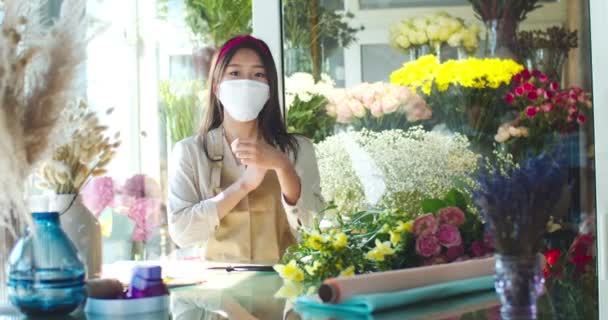 Hermosa mujer asiática florista usando delantal, trabajando en la tienda de flores. Encantadora trabajadora con máscara médica cruzando brazos y mirando a la cámara. Salud, coronavirus, concepto de período pandémico. — Vídeos de Stock