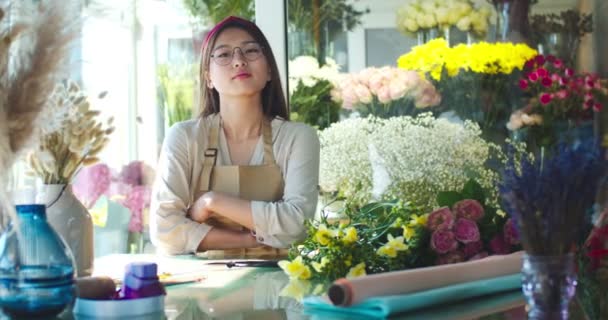 En ung asiatisk kvinnlig ägare som bär förkläde och jobbar i blomsteraffären. Attraktiv kvinnlig florist i glasögon med korsade armar tittar på kameran och ler. Affärs-, butiks- och entreprenörskoncept. — Stockvideo