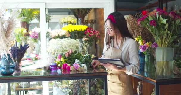 Hardwerkende jonge vrouwelijke winkel eigenaar in schort werken in bloemenwinkel. Mooie Aziatische vrouw bloemist in glazen controleren beschikbaarheid van planten, het nemen van de inventaris op tablet. Bedrijfsleven, ondernemerschap. — Stockvideo