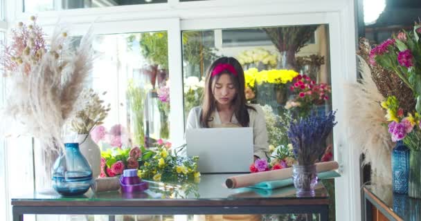 Boa aparência mulher asiática florista vestindo avental, trabalhando na loja de flores. Trabalhadora concentrada usando laptop para receber ordens através da Internet. Negócio, tecnologias, conceito de natureza. — Vídeo de Stock