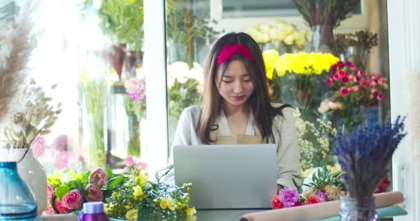 Vacker blomsteraffär ägare varnar förkläde, arbetar i butik. Ganska ung asiatisk kvinnlig florist använder bärbar dator för att chatta med klient, kund, bläddra nyhetsflöde. Sociala medier, teknik, affärsidé. — Stockvideo