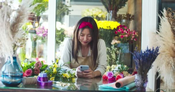 花の店で働くエプロンを身に着けている魅力的な若い店の所有者。美しい女性の花屋は、顧客にメッセージを入力し、スマートフォンで顧客とチャットし、カメラを見て笑っています。事業. — ストック動画