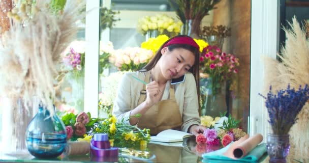 Hermosa joven con delantal, trabajando en la tienda de flores. Florista femenina bastante asiática hablando con el cliente en el teléfono celular y anotando orden de los clientes. Negocios, emprendimiento, concepto de naturaleza. — Vídeos de Stock