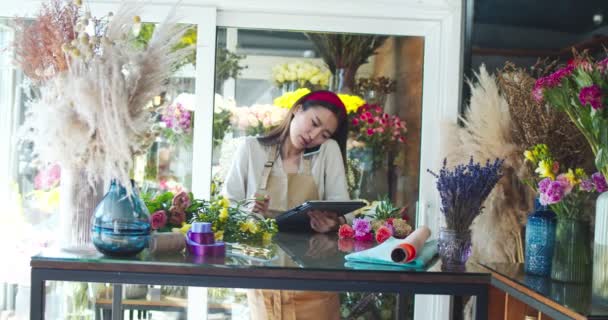 Charmante Aziatische bloemist draagt schort, werkt in een bloemenwinkel. Aantrekkelijke vrouwelijke verkoper die de klant belt, telefoneert met de klant en typt op digitale tablet. Bedrijfs- en technologieconcept. — Stockvideo