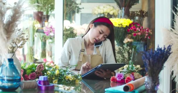 在花店工作的围裙中迷人的年轻女子的画像。英俊的亚洲女花匠打电话给客户，与客户交谈，在平板电脑上打字。商业、技术、小玩艺儿概念. — 图库视频影像
