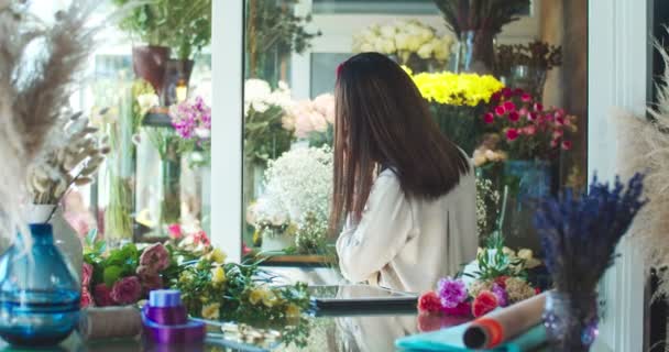 穿着围裙的迷人的亚洲女花匠走到窗前，透过花朵眺望。快乐的年轻女性卖家用智能手机与顾客交谈，用平板电脑定单打字。业务概念. — 图库视频影像