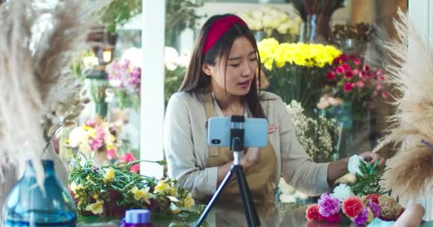 Atractiva mujer asiática florista tiroteo blog para seguidores, espectadores en la tienda de flores. Mujer florista sonriente influenciadora haciendo una conferencia en vivo, oliendo a flor y hablando en cámara. Redes sociales. — Vídeos de Stock