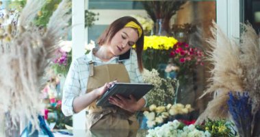 Çiçekçide çalışan önlüklü çekici genç bir kadının portresi. Güzel beyaz kadın çiçekçi müşteriyi arıyor, müşteriyle konuşuyor ve tablet üzerinde yazıyor. İş, teknoloji, aletler konsepti.