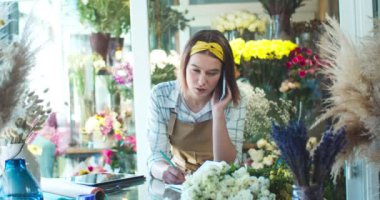 Güzel görünümlü beyaz çiçekçinin portresi çiçekçiden müşteriye sesleniyor. Önlüklü çekici bir bayan işçi müşteriyle konuşuyor ve siparişleri yazıyor. İş, teknoloji, online konsept.