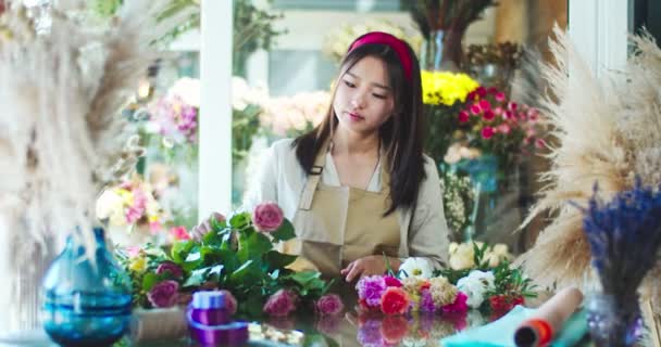 Mujer asiática encantadora usando delantal, trabajando en la tienda de flores. Florista femenina bastante joven que toma flores y corta tallos de plantas con tijeras. Jardinería, naturaleza, concepto de negocio. — Vídeos de Stock