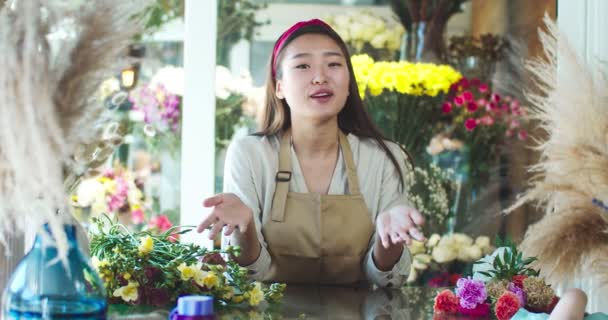 Aantrekkelijke Aziatische vrouw blogger in schort schieten video in bloemenwinkel. Goed uitziende vrouwelijke bloemist maken live conferentie voor volgers, kijkers. Mooie vlogger vertelt over bloemen in de winkel. — Stockvideo