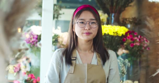 Portret van een knappe Aziatische vrouw in een bril in een bloemenwinkel. Aantrekkelijke jonge vrouwelijke bloemist draagt schort, kijkt en glimlacht naar de camera. Zaken, mensen, natuur concept. — Stockvideo