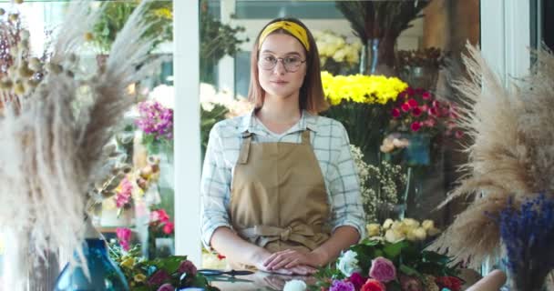 Mooie blanke vrouw bloemist in glazen werken in de bloemenwinkel. Gelukkige jonge werkster met een schort, kijkend en glimlachend naar de camera. Bedrijfsleven, ondernemerschap, beroepsconcept. — Stockvideo