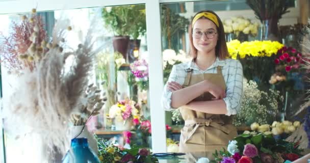 Çiçekçide çalışan önlüklü güzel beyaz kadın dükkan sahibi. Gözlüklü çekici kadın çiçekçi kollarını kavuşturup kameraya bakıyor ve gülümsüyor. İş, girişimcilik kavramı. — Stok video