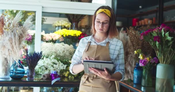 Attraktive kaukasische Blumenverkäuferin in Gläsern, die im Geschäft arbeiten. Charmante junge Floristin tippt auf dem Tablet, schaut in die Kamera und lächelt. Menschen, Berufe, Beruf, Geschäftskonzept. — Stockvideo