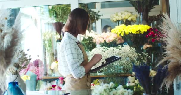 Mooie jonge vrouwelijke bloemist die werkt in een moderne bloemenwinkel. Aantrekkelijke blanke vrouw met schort, inventaris opmaken, laptop gebruiken. Happy shop eigenaar op zoek naar camera en glimlachen. — Stockvideo