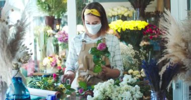 Çiçekçide çalışan önlük giyen çekici genç bir bayan satıcı. Tıbbi maskeli güzel beyaz kadın çiçekçi çiçek alıyor ve gül sapları kesiyor. İş, koronavirüs konsepti.