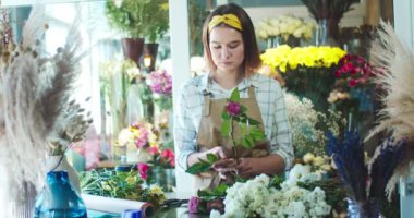Güzel beyaz kadın satıcı önlük giyiyor, çiçekçide çalışıyor. Çiçek alan ve saplarını makasla kesen çalışkan genç bir çiçekçi. Bitkiler, iş, işgal kavramı.