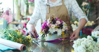 Önlük giyip buket yapan genç beyaz bir çiçekçiyi yakından çek. Çiçek satan güzel bir kadın, şapka kutusuna çiçek takarak mor kurdele bağlıyor. İş, doğa konsepti.
