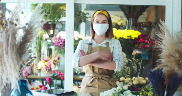 Çiçekçide çalışan, önlük giyen genç, pozitif bir kadın satıcı. Sağlık maskeli güzel beyaz çiçekçi kollarını kavuşturup kameraya bakıyor. İş, meslek, doğa kavramı. — Stok video