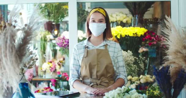 Snygg kvinnlig florist bär förkläde, arbetar i modern blomsteraffär. Fattiga unga kaukasiska visa ägare i medicinsk mask tittar till kameran. Pandemisk period, säkerhetsåtgärder, hälso- och sjukvård. — Stockvideo