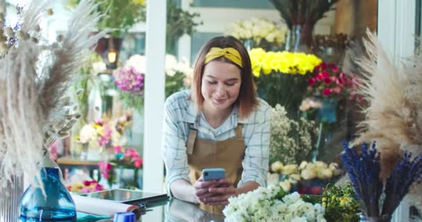 Bella giovane lavoratrice in grembiule che lavora nel negozio di fiori. Affascinante donna caucasica fiorista digitando, inviando un messaggio ad un amico su smartphone e sorridente. Social media, Internet. — Video Stock