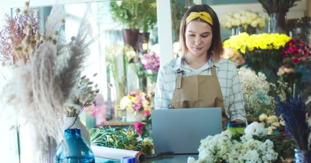 Goed uitziende blanke bloemenwinkel eigenaar in schort werken in de winkel. Mooie jonge vrouwelijke bloemist met behulp van laptop, typen, e-mails controleren en koffie drinken. Bedrijfsleven, technologie, internetconcept. — Stockvideo