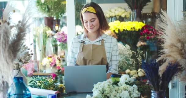 Linda florista feminina caucasiana em avental trabalhando em laptop na loja de flores. Proprietário da loja mulher atraente batendo ordens on-line, e-mails, e mensagens de digitação. E-commerce, negócio, conceito de Internet. — Vídeo de Stock