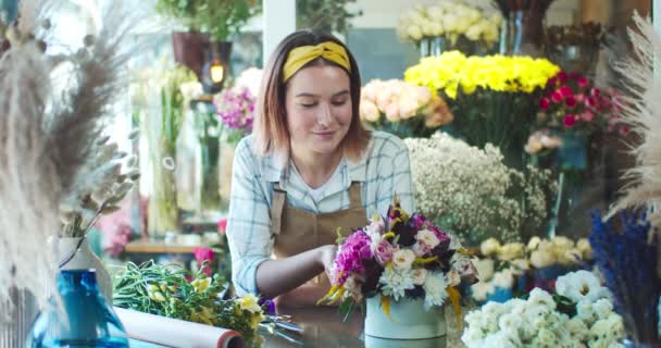Mooie jonge bloemenwinkel eigenaar draagt schort, werken in de winkel. Charmante blanke vrouwelijke bloemist die over het boeket waakt, kijkt en lacht naar de camera. Bedrijfs- en ondernemerschapsconcept. — Stockvideo