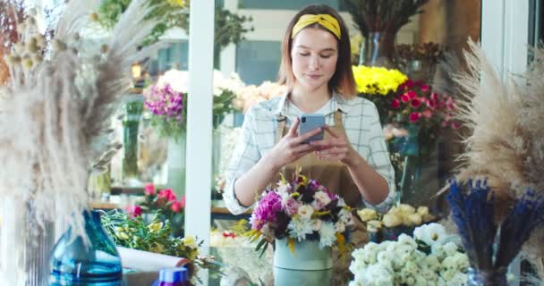 Vrolijke blanke bloemenwinkel verkoper draagt schort, werkt in de winkel. Mooie jonge vrouwelijke bloemist neemt foto voor de klant en stuurt foto 's naar de klant. E-commerce, business, online concept. — Stockvideo