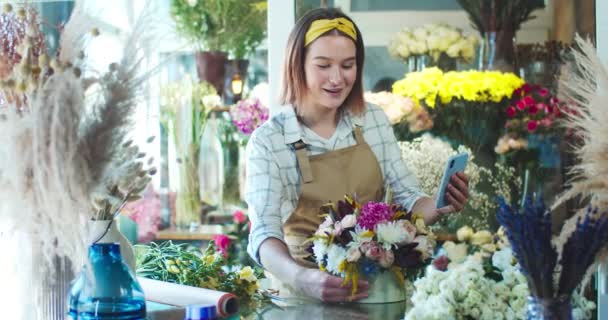 Goed uitziende blanke winkelier praat met Cleint via video face chat in de bloemenwinkel. Gelukkige jonge vrouwelijke bloemist bellen naar de klant om boeket van bloesems te tonen. Bedrijfsconcept, social media. — Stockvideo