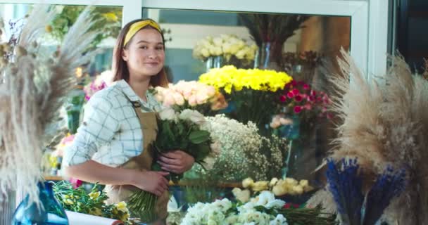 Linda florista mulher caucasiana que trabalha na loja de flores moderna. Trabalhadora encantadora vestindo avental, segurando buquê de rosas, olhando e sorrindo para a câmera. Negócio, conceito de ocupação. — Vídeo de Stock