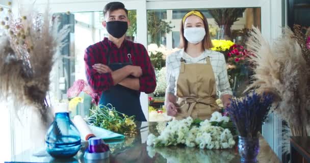 Jonge mannelijke en vrouwelijke bloemisten in medische maskers dragen schort in bloemenwinkel. Blanke man en vrouw arbeiders kruisen armen en kijken naar de camera. Bedrijfs- en handelsconcept. Coronavirus, pandemie. — Stockvideo