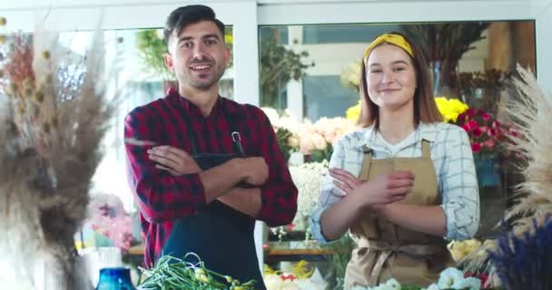 Enthousiaste mannelijke en vrouwelijke bloemisten dragen een werkschort in de bloemenwinkel. Jonge blanke vrouwen en mannen kruisen hun armen, kijken naar de camera en glimlachen. Zakelijke en commerciële betrekkingen. — Stockvideo