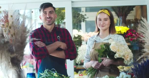 エプロンを着た白人女性と男性の花屋は、カメラを見て、花の店で笑顔。ハンサムな男が腕を渡り、美しい女性が花束を持っています。ビジネス、商取引の概念. — ストック動画